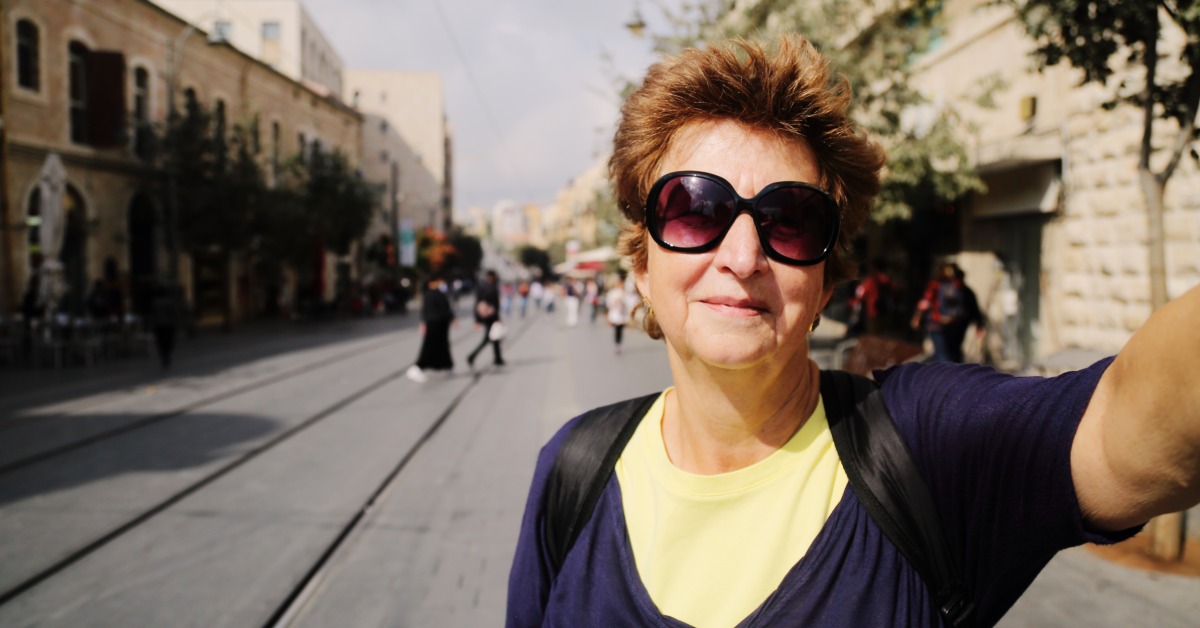A senior woman in sunglasses takes a selfie on an Israel street. She is on a multigenerational tour of the country.