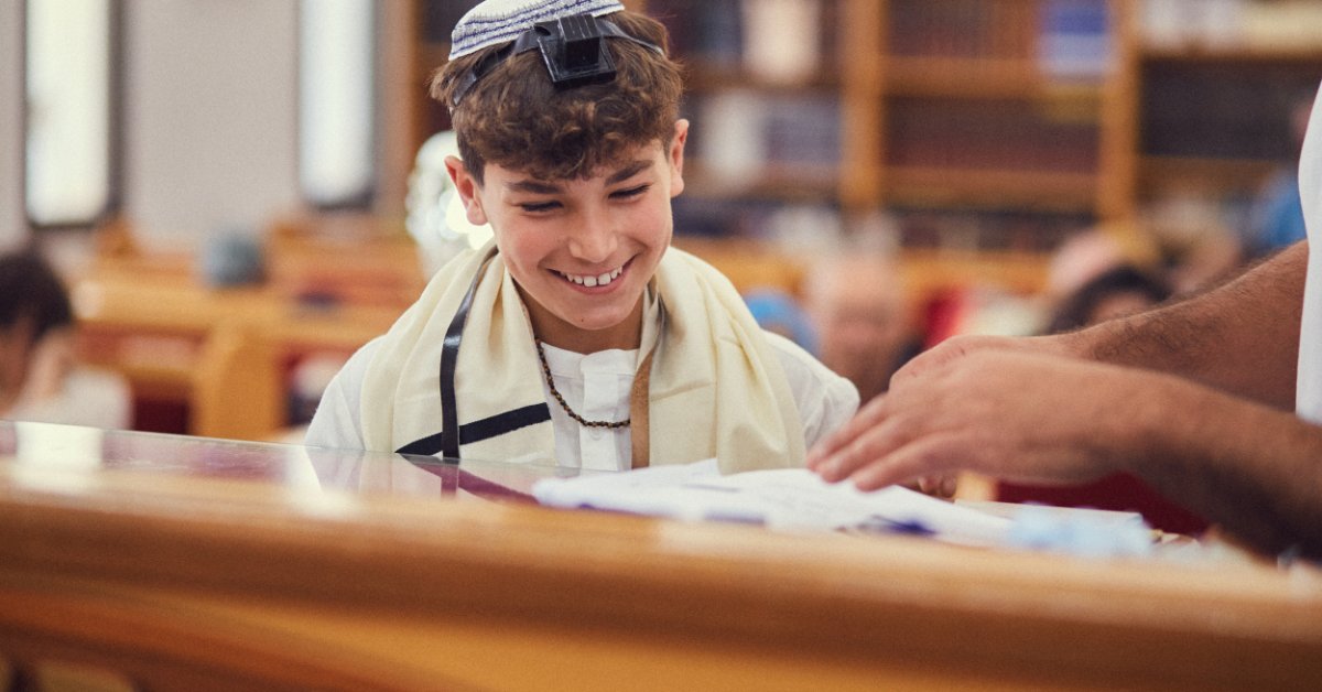 A young Jewish boy at his Bar Mitzvah, smiling as he stands with a member of the ceremony. He wears a yamaka.