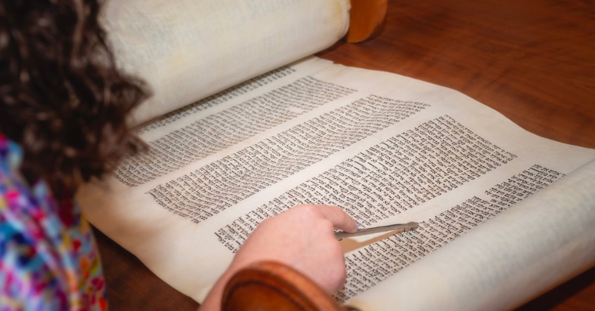 A young Jewish girl's hand scrolls the text in the Torah at her Bat Mitzvah. She uses a pointer to follow the words.